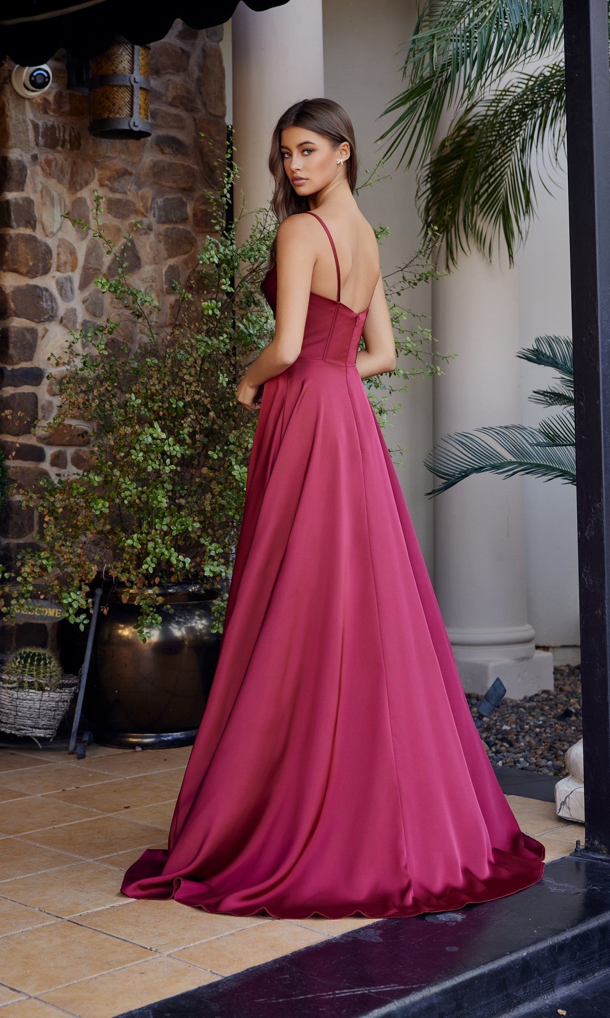 A woman in a long, sleeveless magenta Nox Anabel A1744 prom dress stands on a patio with stone walls, lush plants, and elegant columns in the background.